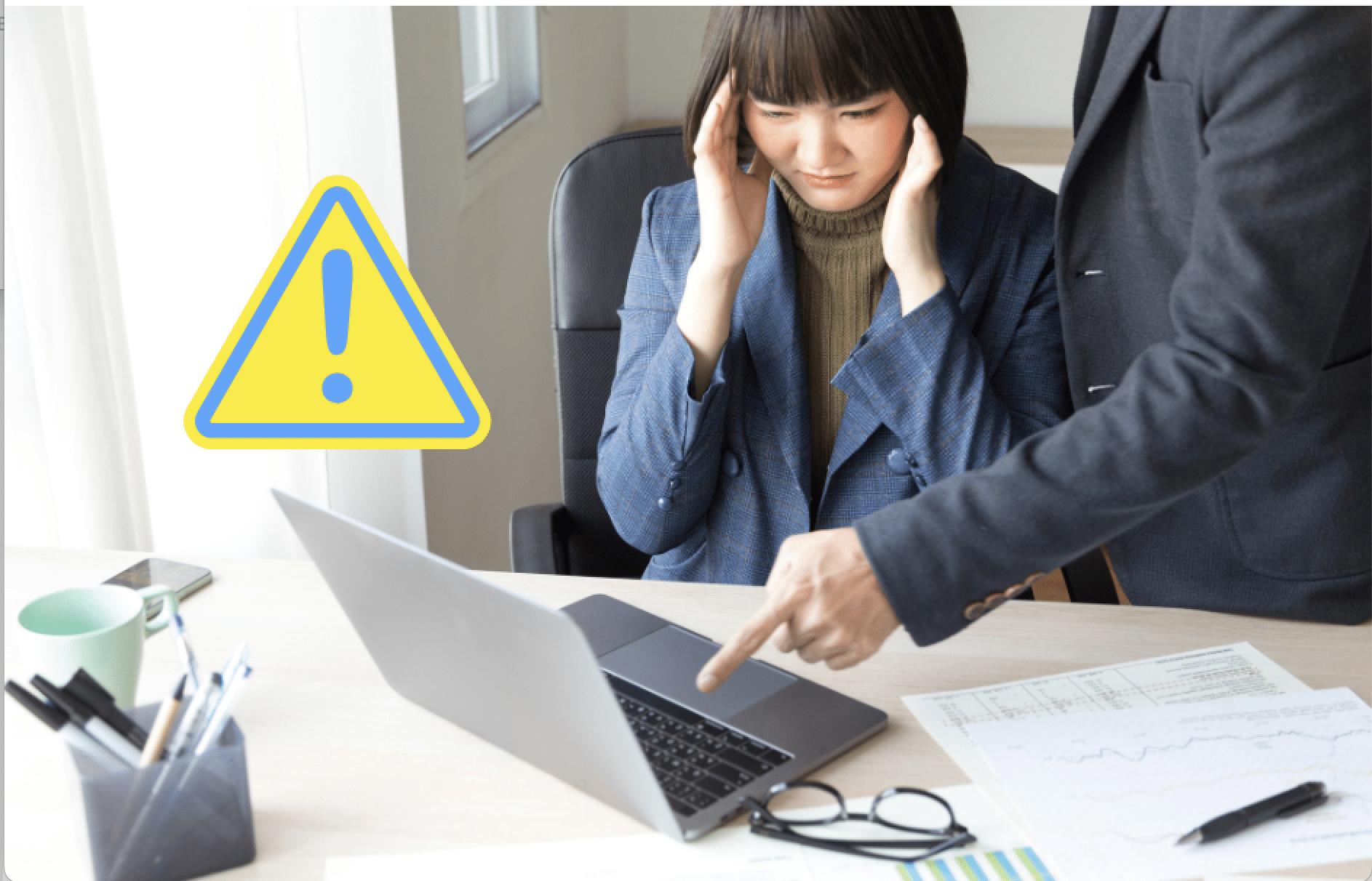 Woman looking stressed at a laptop while a colleague points at the screen, with a warning symbol, illustrating poor communication and workplace conflict.